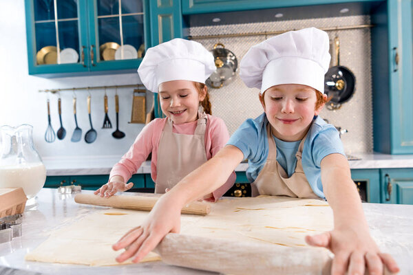 happy little kids in chef hats and aprons rolling dough in kitchen