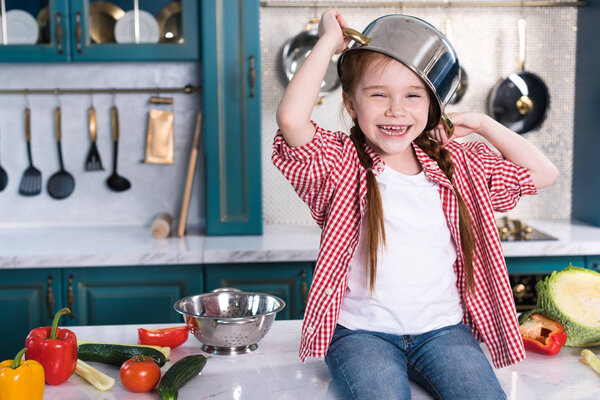 cute kid with pan on head having fun and sitting on kitchen table