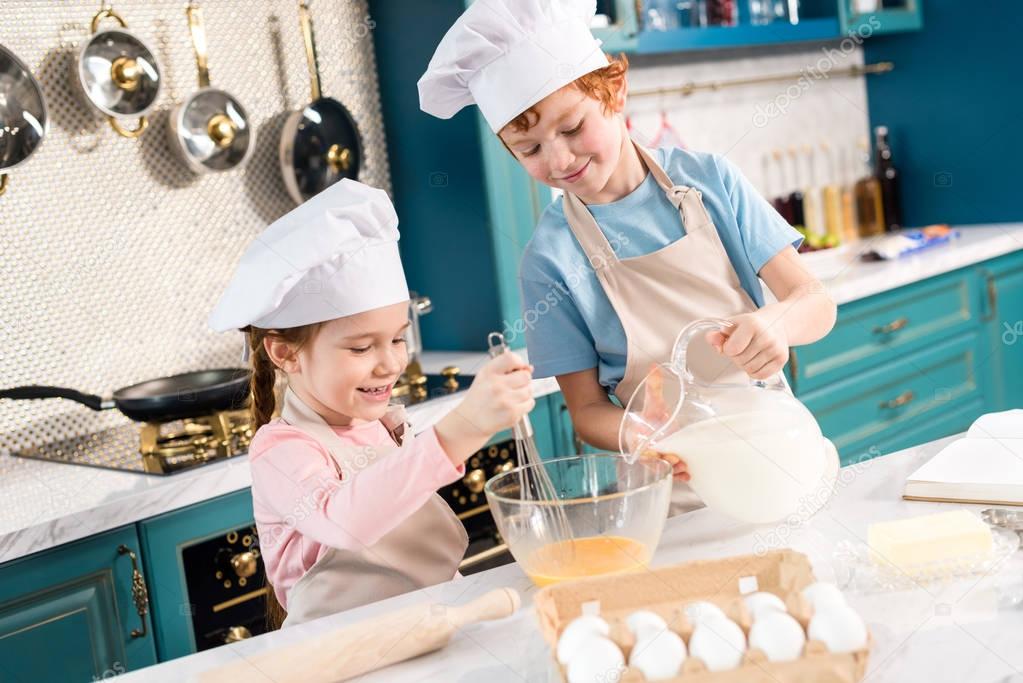 happy little kids in chef hats and aprons preparing dough together in kitchen