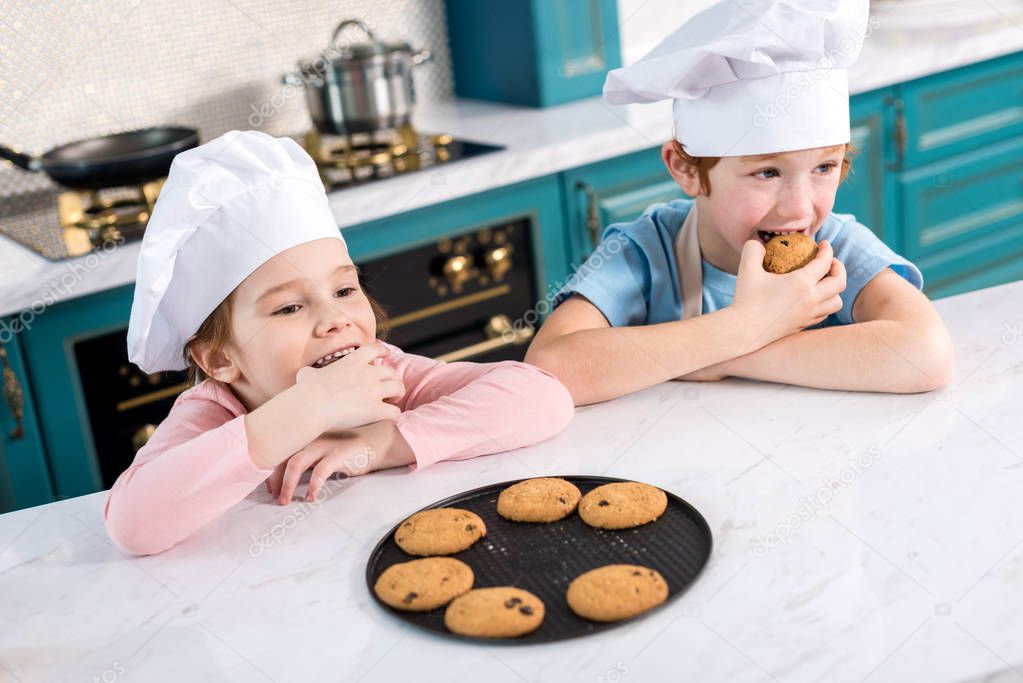happy children in chef hats eating tasty cookies in kitchen