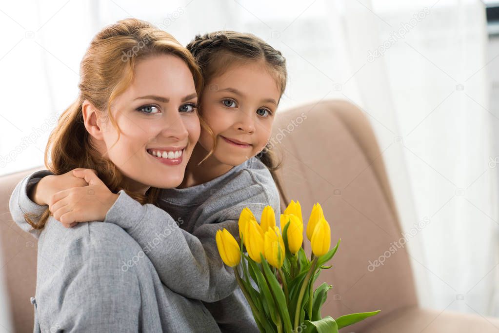 happy mother and daughter with yellow tulips hugging and smiling at camera