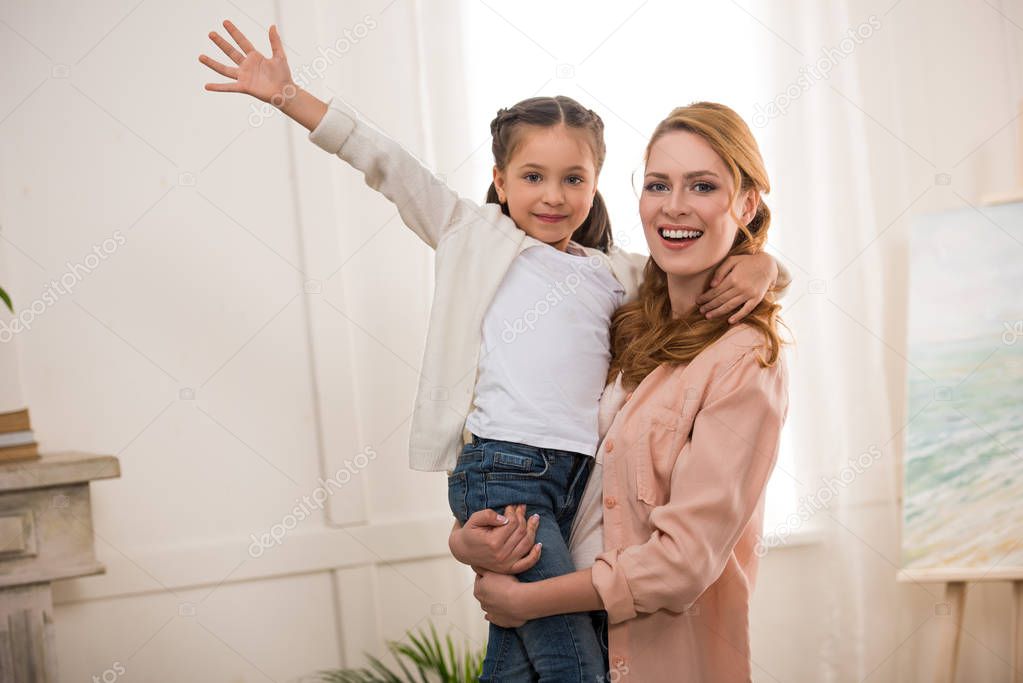young woman carrying adorable little daughter and smiling at camera at home