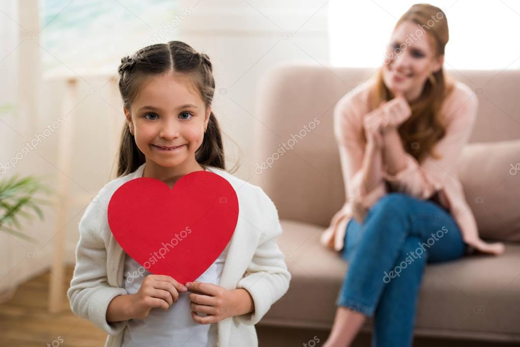 adorable child holding red heart symbol and smiling at camera while mother sitting behind