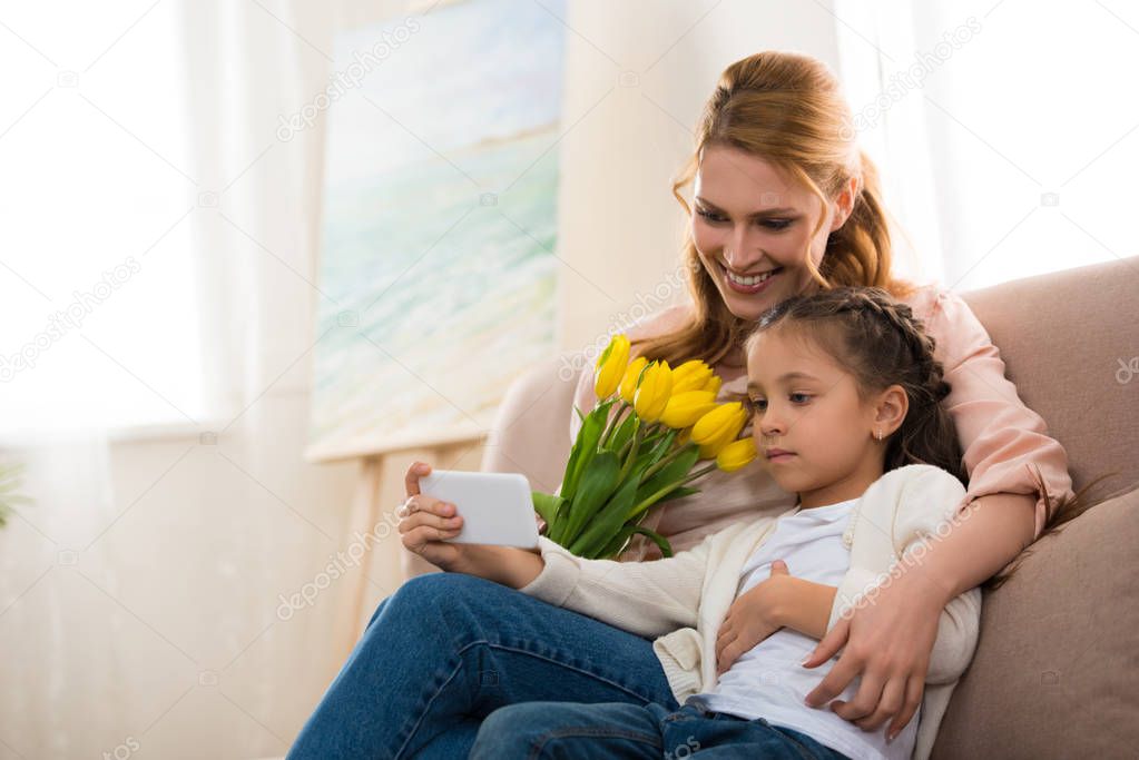 happy mother and daughter with yellow tulips using smartphone together at home