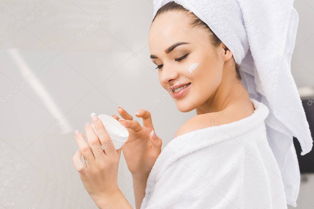 side view of smiling beautiful woman in bathrobe with towel on head holding face cream