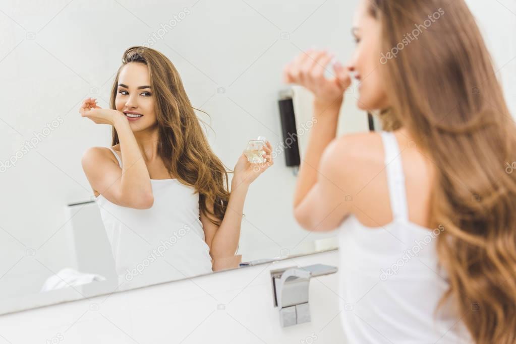 mirror reflection of smiling woman smelling perfume on hand in bathroom