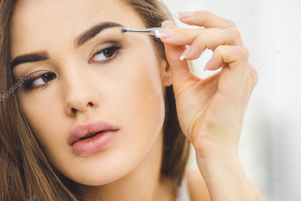 portrait of young woman plucking eyebrows with tweezers