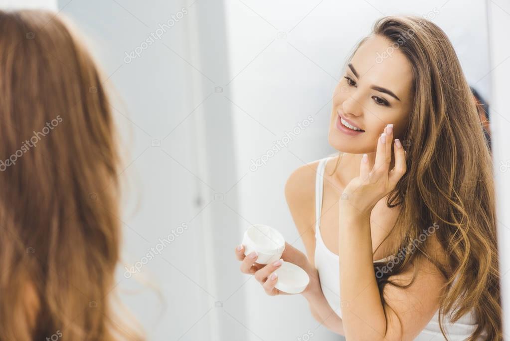 mirror reflection of beautiful smiling woman applying face cream