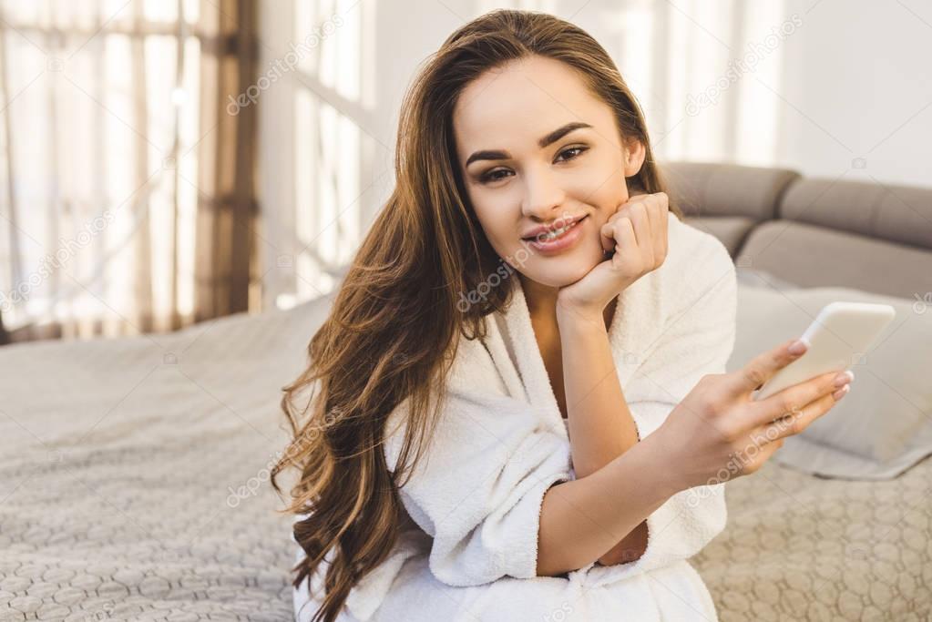 smiling young woman in bathrobe with smartphone sitting on bed