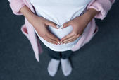 oříznutý pohled těhotná dívka dělat symbol srdce s rukama na břiše