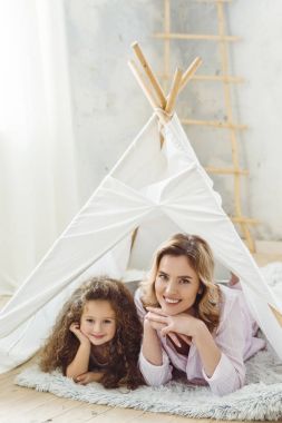 beautiful cheerful mother and curly daughter lying in kid wigwam clipart