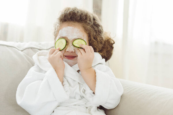 child in bath robe with cucumber slices on eyes and facial mask on face