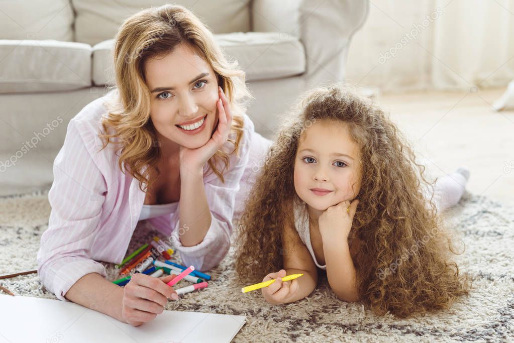 curly daughter and beautiful mother with markers and drawing album looking at camera