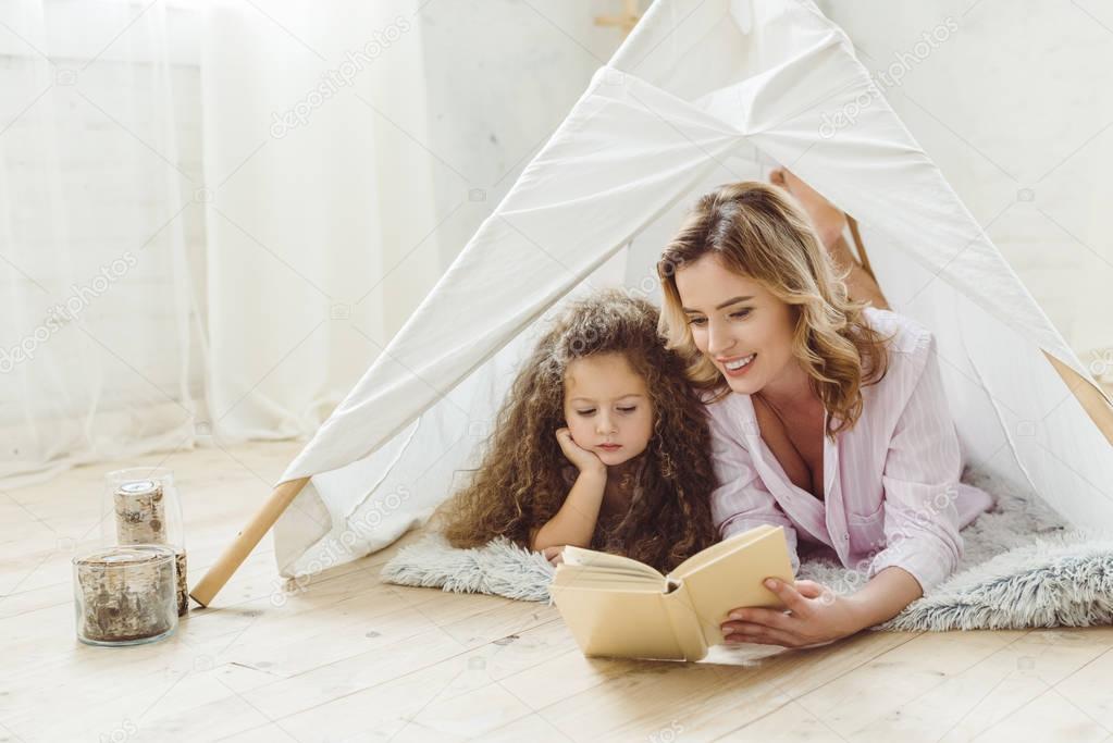 happy mother and daughter reading book together in kid wigwam