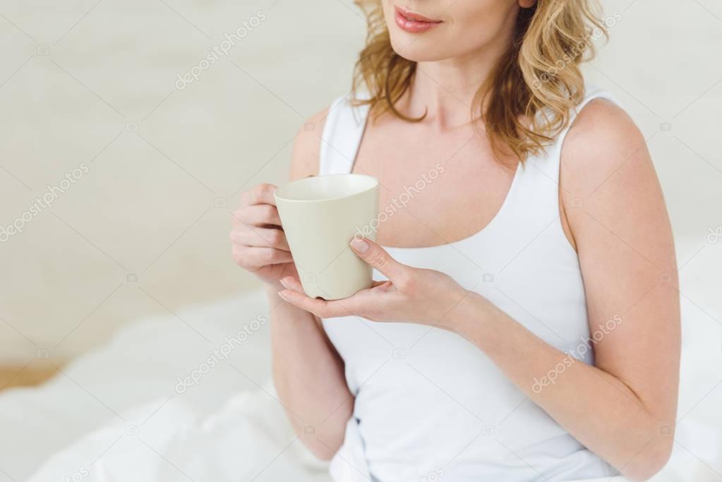 cropped view of woman with cup of coffee in bedroom in the morning