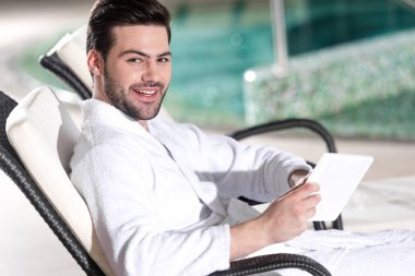 Dijital tablet tutarak ve spa merkezi kamerada gülümseyen bornoz yakışıklı adam 