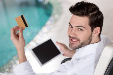 kredi kartı ve dijital tablet spa merkezinde tutarak bornoz gülümseyen genç adam   
