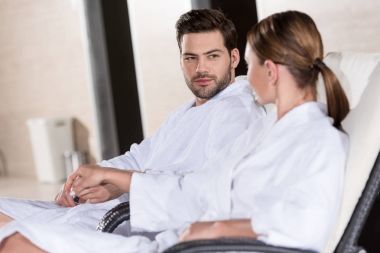 young couple in bathrobes holding hands and looking at each other while resting together in spa center