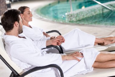 spa merkezi havuza yakın oturan bornoz mutlu genç çift yan görünüm