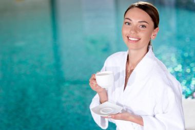 genç kadın Kupası tutarak ve spa merkezi kamerada gülümseyen bornoz