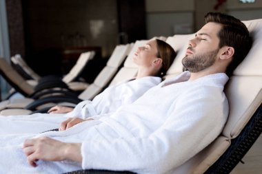side view of young couple in bathrobes resting together in spa center clipart