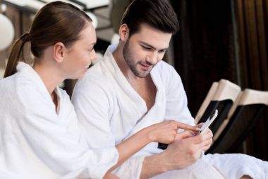 Genç iki smartphone birlikte spa Center'da kullanarak bornoz içinde gülümseyen 
