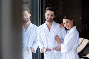 happy young couple in bathrobes smiling at camera in spa center clipart