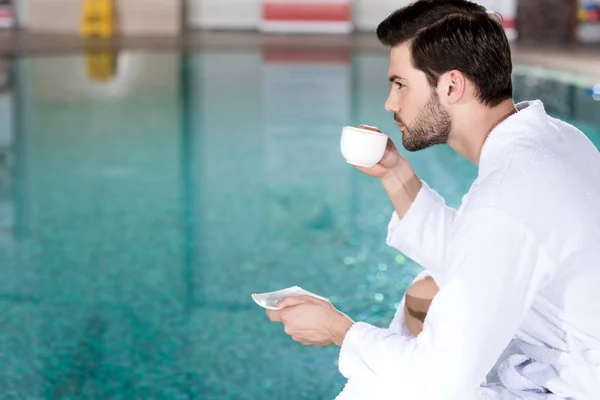 ウェルネス センターのプールのそばに座ってコーヒーを飲みながらバスローブの男の側面図 — ストック写真