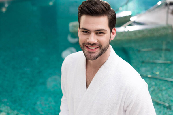 handsome young man in bathrobe smiling at camera while standing in spa center