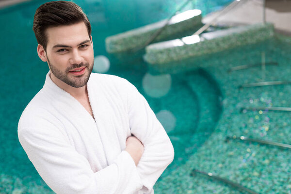 handsome young man in bathrobe standing with crossed arms and looking at camera in spa center