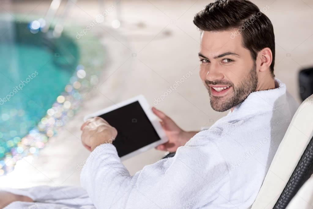 handsome young man in bathrobe holding digital tablet and smiling at camera in spa center 