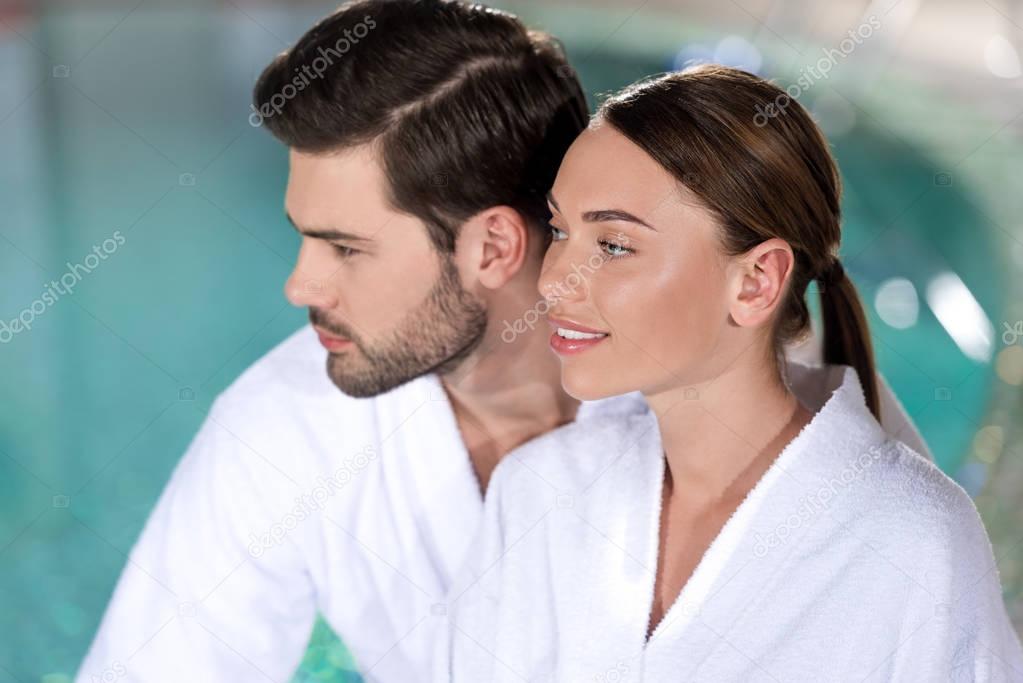 smiling young couple in bathrobes looking away in spa center