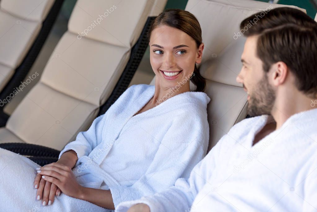 happy young couple in bathrobes smiling each other while resting together in spa center