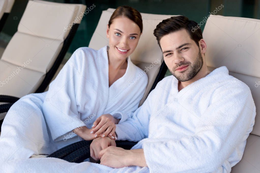 happy young couple in bathrobes smiling and camera in spa center