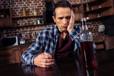 upset man sitting at table with bottle and glass of alcohol at home clipart