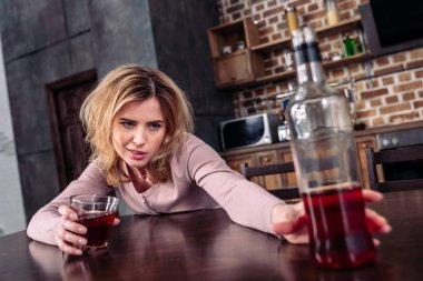 selective focus of drunk woman taking bottle of alcohol while sitting at table at home clipart