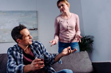 man with glass of alcohol sitting on sofa while arguing with wife behind at home clipart