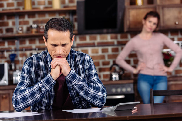selective focus of pensive man sitting at table and wife standing behind at home, financial problems concept