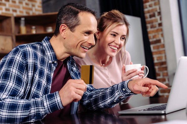 happy couple with credit card and cup of coffee looking at laptop screen at home