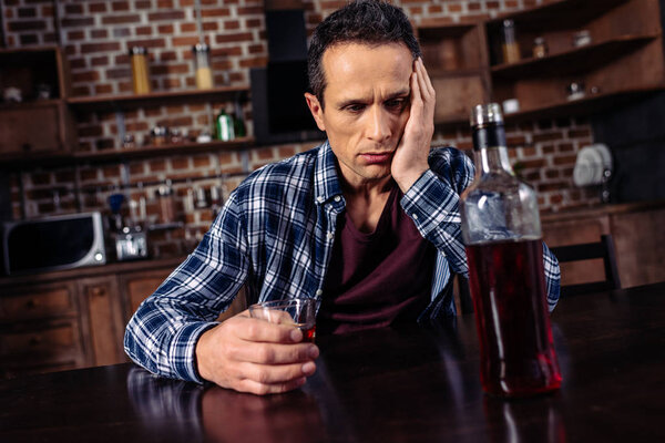расстроенный мужчина сидит за столом с бутылкой и стаканом алкоголя дома
