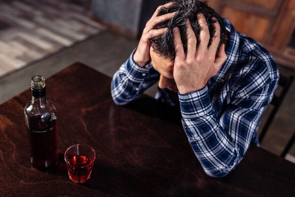 высокий угол обзора пьяного человека, сидящего за столом с бутылкой и стаканом алкоголя дома
