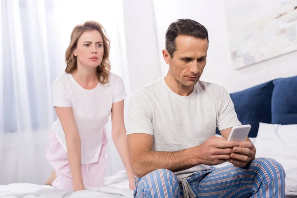 困惑的妻子看着丈夫使用智能手机 而坐在家里的床上 — 图库照片