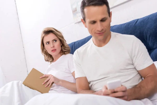 Foco Seletivo Homem Usando Smartphone Enquanto Esposa Ler Livro Cama — Fotografia de Stock