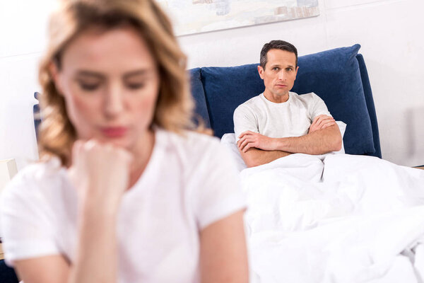 selective focus of upset woman sitting on bed with husband behind at home, relationship difficulties concept