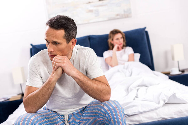 selective focus of upset man sitting on bed with wife behind at home, relationship difficulties concept