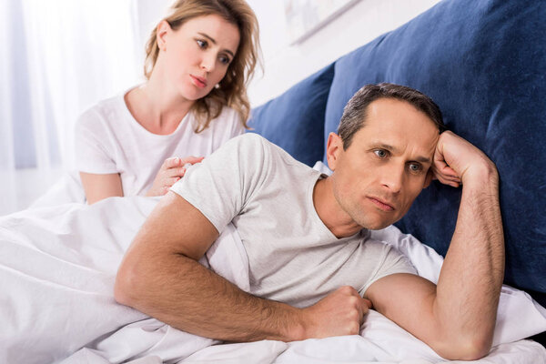 portrait of upset man lying in bed with wife behind