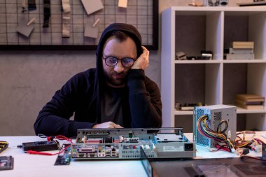 Kapalı gözlerle oturan kapüşonlu adam ve karşı masada kırık pc başına el