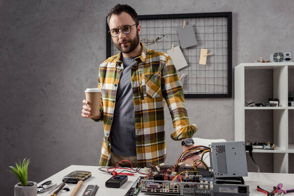 man with coffee in hands standing against table with computer parts 