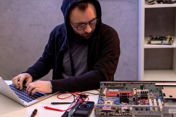 man in hoodie using laptop while looking on broken pc