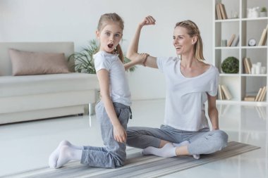 mother showing biceps muscle to her shocked daughter at home